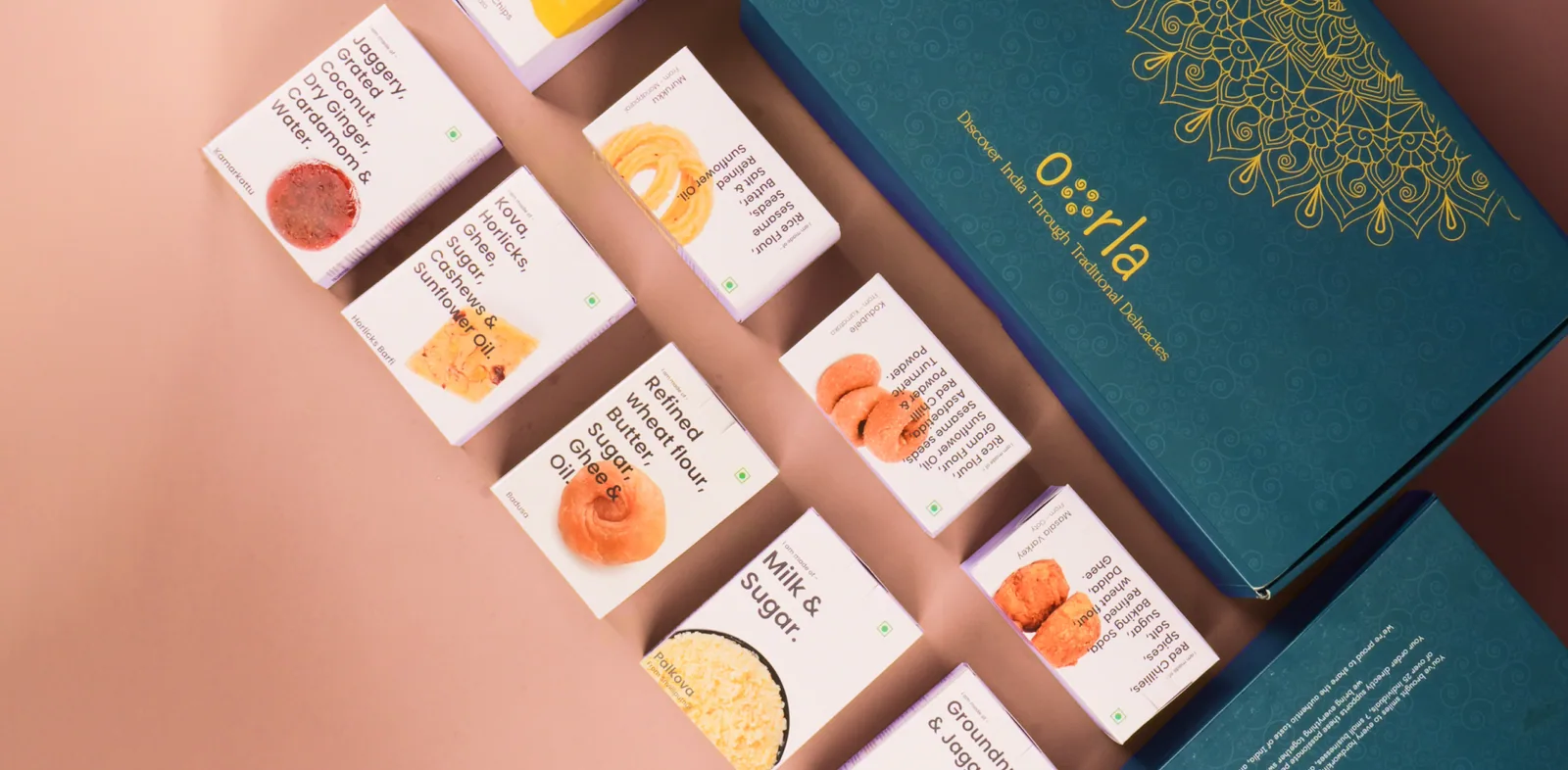 Printed sweet boxes fanned across a pink table, each panel naming its own ingredients, beside a teal Oorla gift box with gold mandala artwork.
