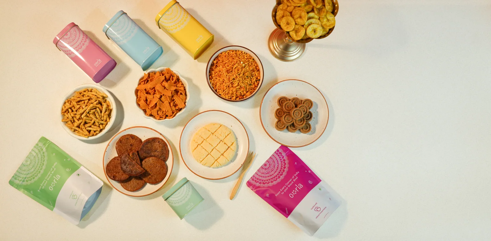 An Oorla order laid out on a white table — pastel printed tins, plates of sweets, bowls of savoury mixture, and printed pouches