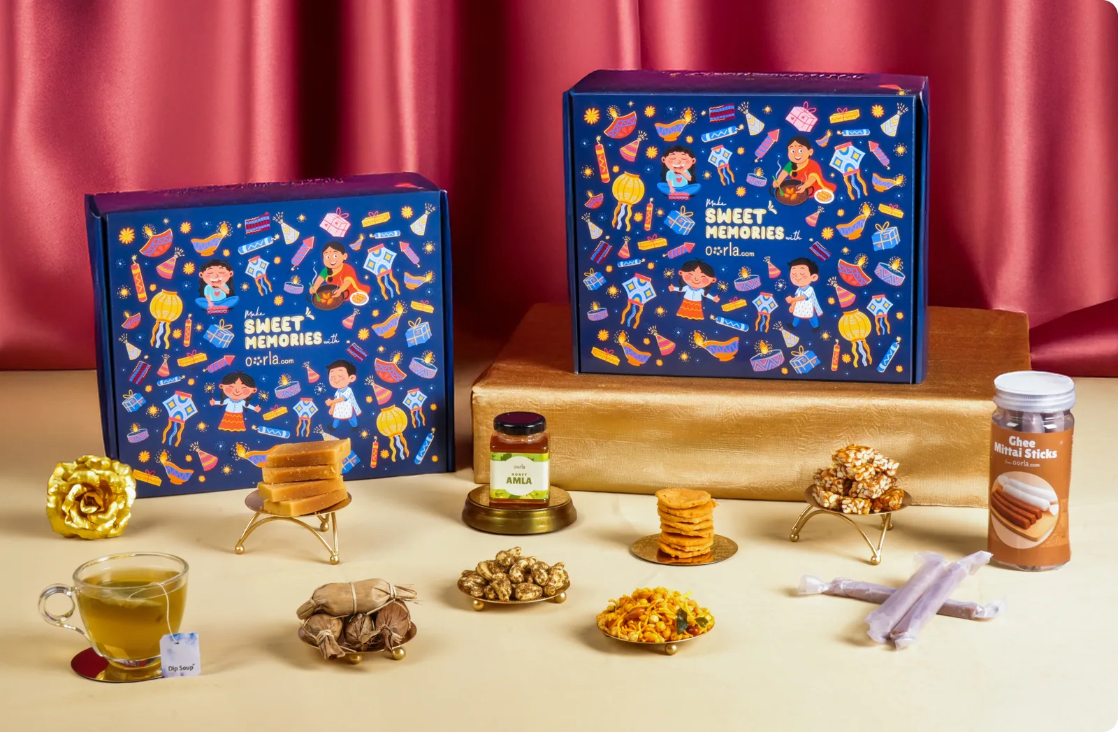 Two illustrated navy Grand Diwali Hamper boxes on a gold plinth against red drapes, with sweets and savouries on brass stands, a jar of amla, ghee mittai sticks and a glass of tea arranged in front
