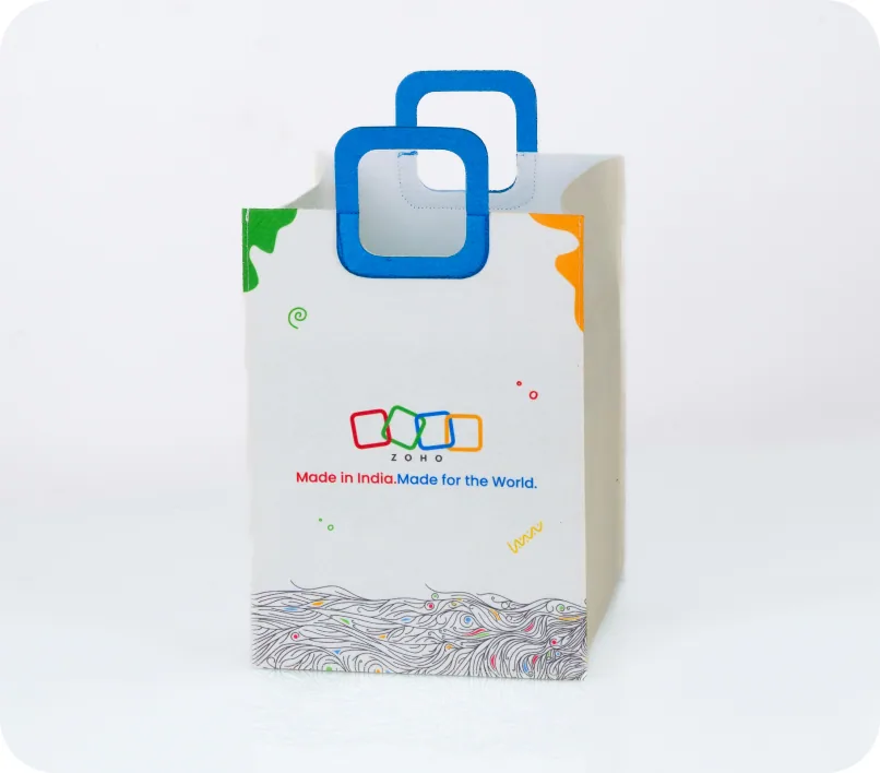 A white gift bag with blue die-cut handles, printed with the ZOHO logo and the line Made in India, Made for the World