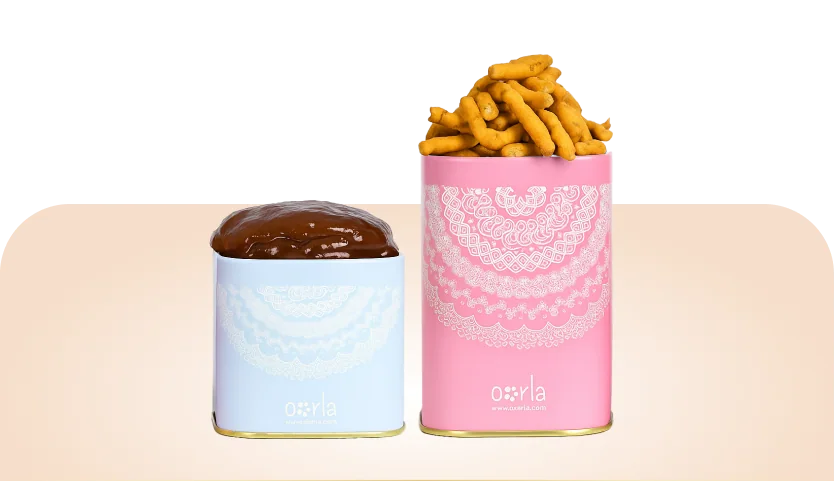 Premium Colourful Tins: a pale blue square tin and a pink cylindrical tin, both printed with white lace patterns and the Oorla mark, filled with halwa and murukku.