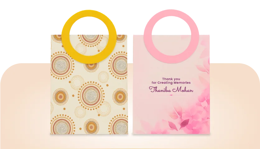 Sustainable Bags: two recycled-board gift bags with die-cut circular handles, one printed with ochre mandalas and one personalised with a recipient's name in pink.