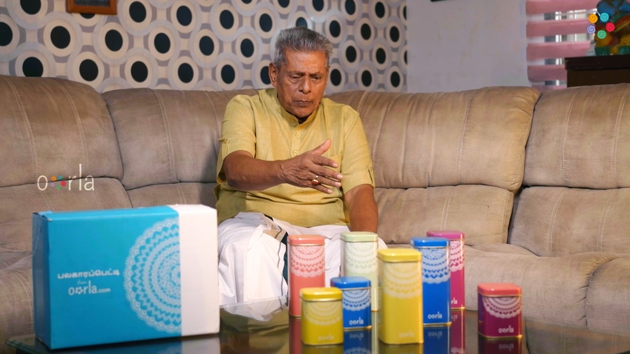 A still from the film: an elderly man in a yellow kurta sitting at home, talking across a table laid with an Oorla sweet box and a row of printed snack tins.